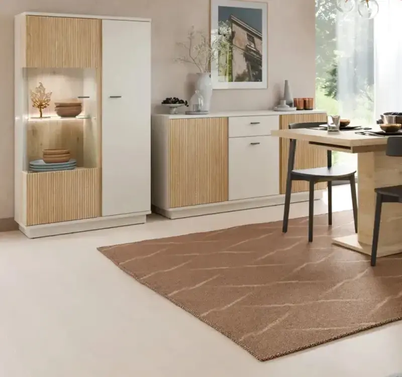 Esszimmer mit weisser Vitrine und passendem Sideboard mit Holzlamellen, darauf Vasen und Geschirr, vorn rechteckiger Esstisch mit Stühlen auf braunem Teppich vor Fenster.